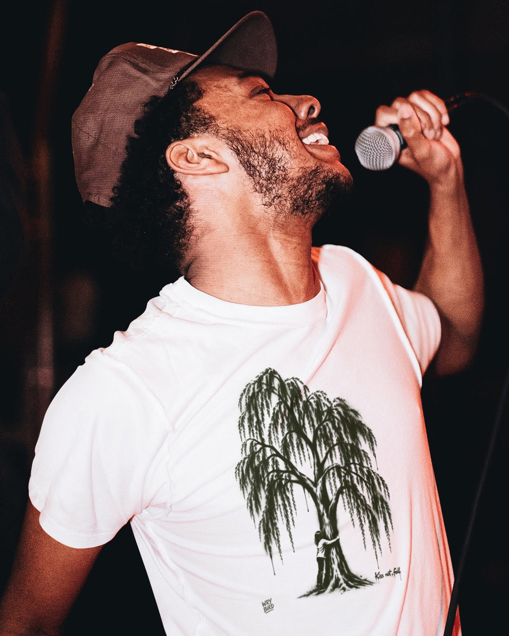 A man wearing a white t-shirt with a green willow tree graphic and a brown cap, singing into a microphone on stage against a dark background. By Wry Bird.