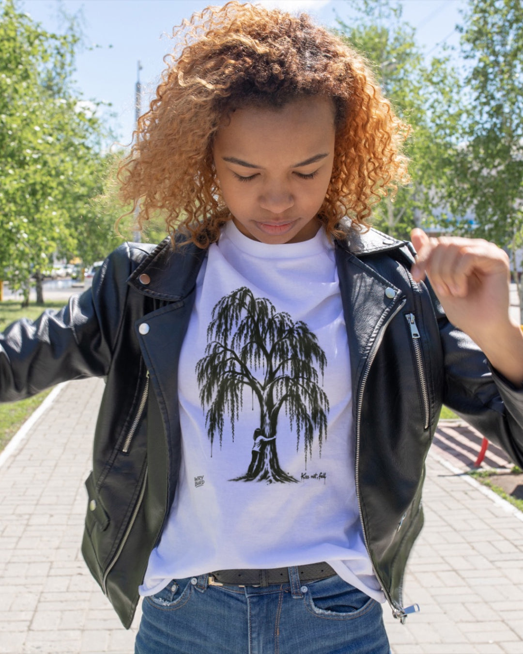 A woman wearing a white graphic t-shirt with a Wry Bird weeping tree print, styled with a black leather jacket and blue jeans, standing outdoors on a paved path with trees in the background. Kiss Not Fell