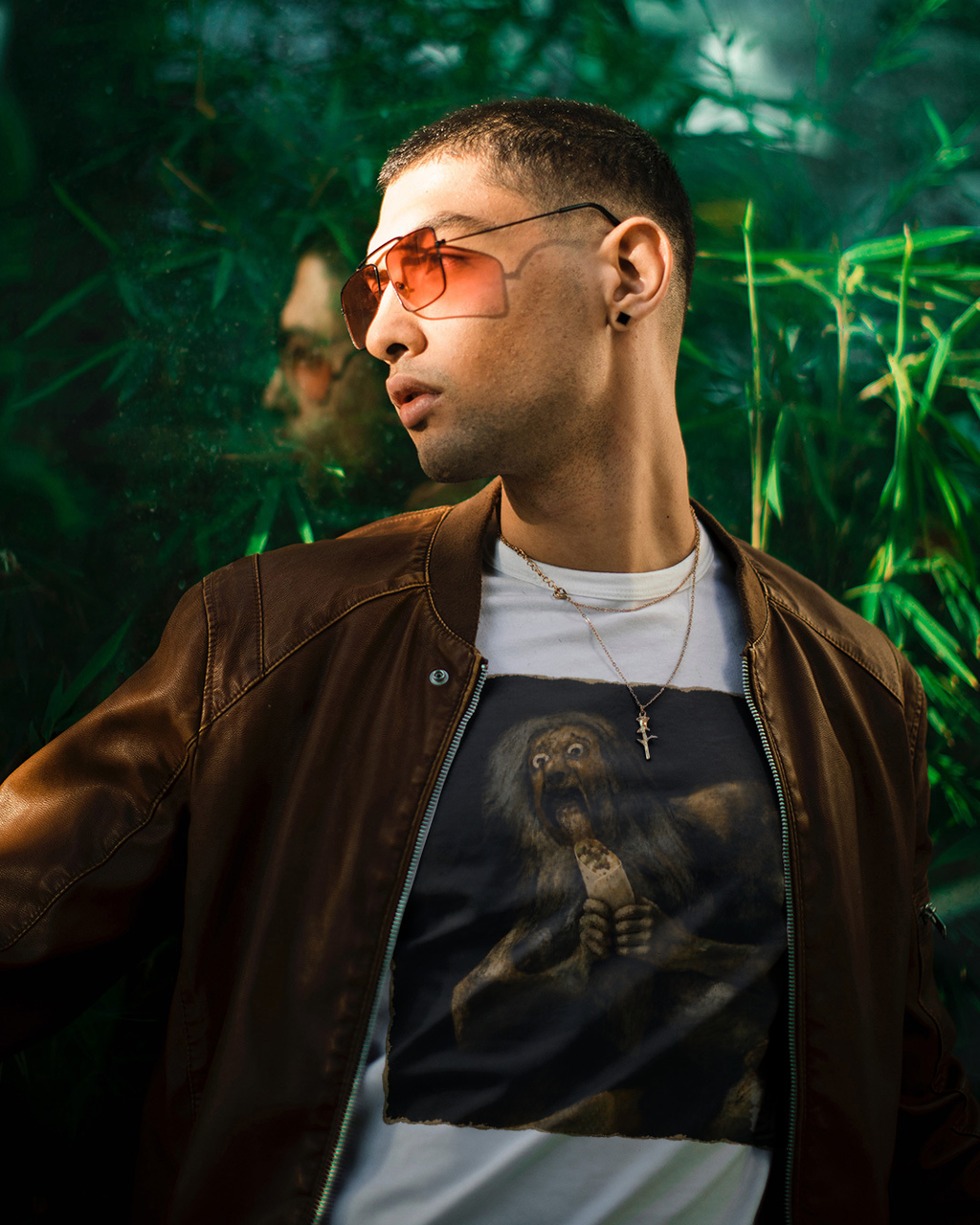 A man wearing orange-tinted aviator sunglasses, a brown leather jacket, and a white t-shirt with a Wry Bird graphic print, set against a dark green foliage background.