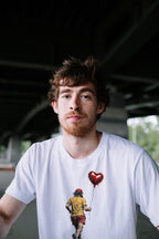 A man wearing a white graphic t-shirt with a red heart balloon and a Wry Bird running figure print, photographed under a bridge.