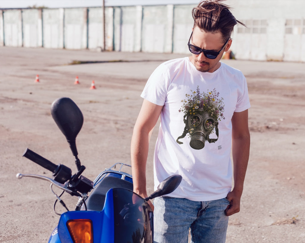 A man wearing a white Wry Bird graphic t-shirt with a gas mask and flowers design, blue jeans, and sunglasses, standing next to a blue motorcycle in an outdoor paved area.