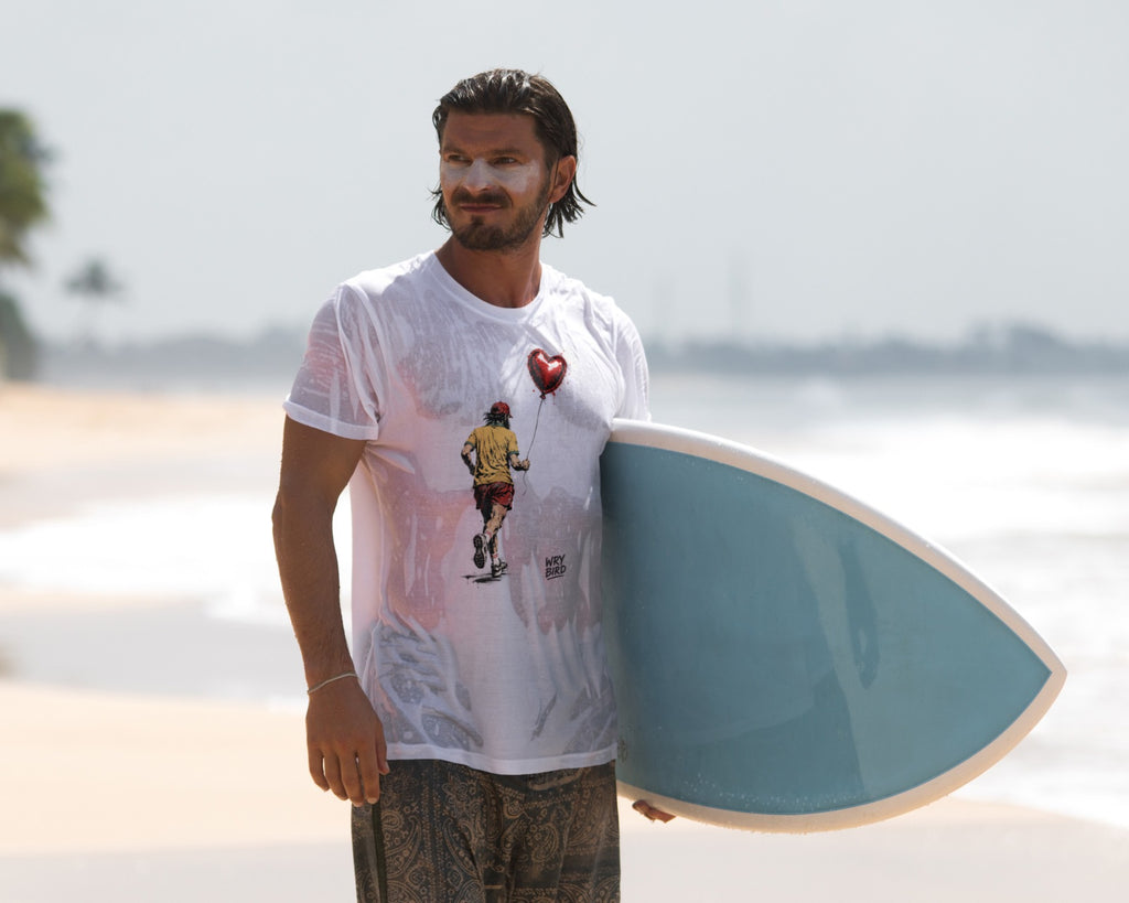 A man standing on a beach holding a light blue surfboard, wearing a white graphic t-shirt with a Wry Bird Banksy style design and patterned shorts.