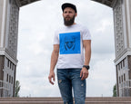 A man wearing a white Wry Bird t-shirt with a blue graphic print of a Saxon helmeted face, paired with distressed blue jeans and a black cap, standing in front of a large stone archway under a cloudy sky.