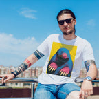 A man wearing a white graphic t-shirt with an Andy Warhol style Wry Bird print, blue denim shorts, and sunglasses, sitting outdoors against a blue sky background.