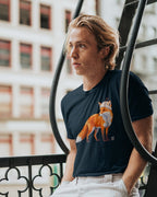 A man wearing a navy blue Wry Bird t-shirt with a graphic of an orange fox wearing a small hat, standing on a black metal fire escape with a blurred city building in the background.