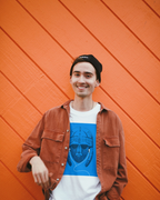 A smiling young man wearing a black beanie, a brown corduroy jacket, and a white t-shirt with a blue Wry Bird Saxon graphic print, standing against a bright orange wood-paneled wall.