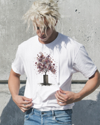 A man with messy platinum blonde hair wearing a white Wry Bird graphic t-shirt featuring a tree with purple and pink foliage growing out of a dark barrel, paired with blue jeans, standing against a textured concrete wall.