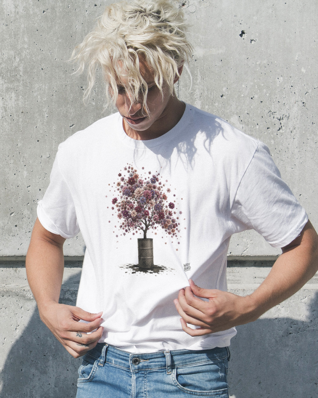 A man with messy platinum blonde hair wearing a white Wry Bird graphic t-shirt featuring a tree with purple and pink foliage growing out of a dark barrel, paired with blue jeans, standing against a textured concrete wall.