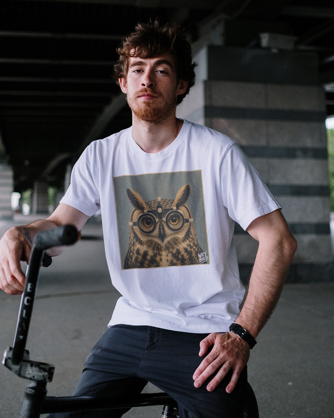 A man wearing a white crewneck Wry Bird t-shirt with a graphic print of an owl wearing round glasses, sitting on a scooter in an outdoor underpass.