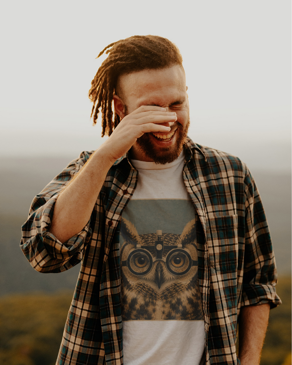 A man with reddish-brown hair and a beard, wearing a green and brown plaid flannel shirt over a white Wry Bird t-shirt with a large owl graphic, laughing outdoors against a soft-focus background.