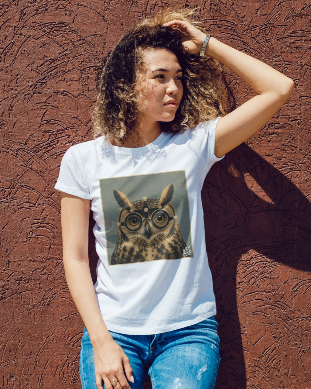 A woman with curly hair wearing a white graphic Wry Bird t-shirt with a square owl print, paired with blue jeans, posing against a textured reddish-brown stucco wall.